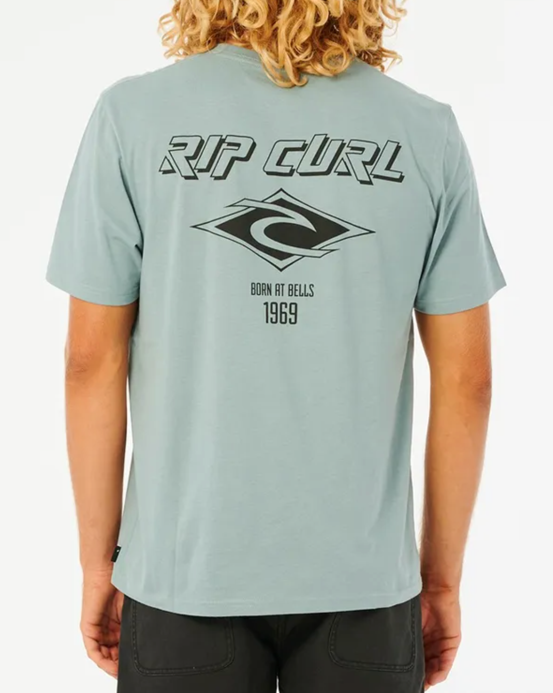 A person with curly hair wears a light blue Rip Curl Fade Out Icon Tee, made from organic cotton, featuring a graphic logo and "BORN AT BELLS 1969" text on the back. The relaxed fit complements dark pants for a casual look.