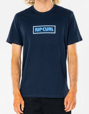 Standing out against the plain white background, a person exudes a laid-back vibe in the Rip Curl Big Mumma Icon Tee—a casual navy blue shirt with light blue "RIP CURL" lettering on the chest. Curly hair cascading down complements the effortless, authentic style.