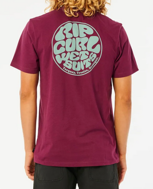 A person with wavy hair is wearing the Rip Curl Wetsuit Icon Tee, a 100% cotton maroon t-shirt featuring "Rip Curl Wetsuits" in large, stylized bubble letters within a circle on the back, alongside the text "Torquay, Trestles, Hossegor." They are also dressed in black pants.