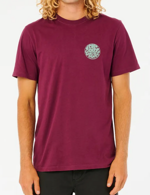 A person with shoulder-length curly blonde hair is wearing a Rip Curl Wetsuit Icon Tee, a short-sleeved maroon T-shirt made of 100% cotton, paired with black pants. The comfortable tee features a small circular Rip Curl print logo on the left chest area. The background is plain white.