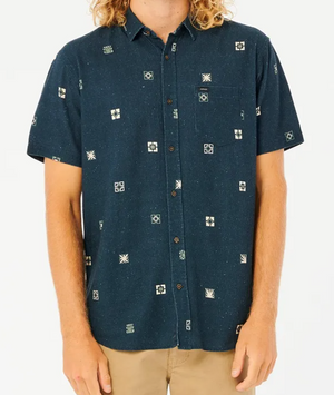 A person models a Rip Curl Party Pack Short Sleeve Shirt in dark blue with geometric patterns against a white background, paired with comfortable light khaki standard-fit pants. Their face is not visible.