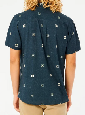 Rear view of a person with curly blonde hair in a dark blue Rip Curl Party Pack Short Sleeve Shirt with small geometric patterns and beige pants, standing comfortably against a white background.