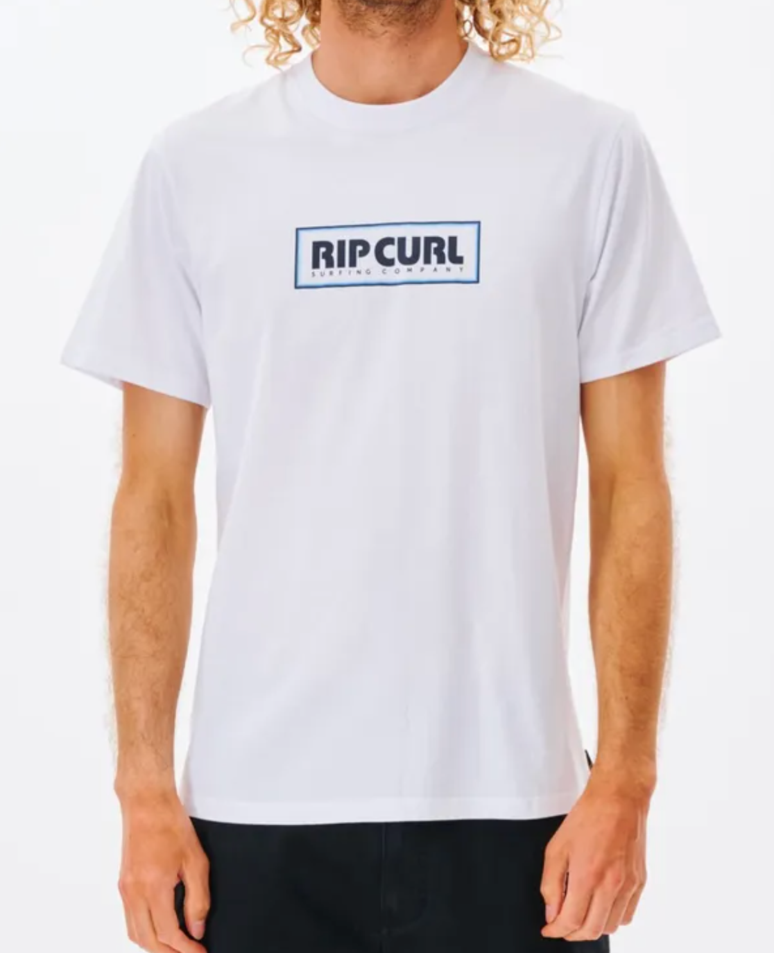 A person with curly hair models the Rip Curl Big Mumma Icon Tee, made from Organic Cotton, showcasing "RIP CURL" in a black box on the chest. They pair it effortlessly with dark pants against a plain white background.