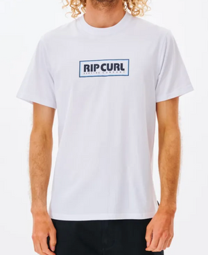 A person with curly hair models the Rip Curl Big Mumma Icon Tee, made from Organic Cotton, showcasing "RIP CURL" in a black box on the chest. They pair it effortlessly with dark pants against a plain white background.