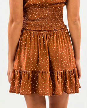 A person is seen from the back wearing a Rip Curl Dreamer Spot Mini Skirt, showcasing its relaxed style with a smocked waist, white polka dots, and ruffled hem. They pair it with a sleeveless brown dress as their arms rest casually at their sides.