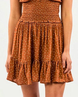 A person wearing a Rip Curl Dreamer Spot Mini Skirt in brown with white polka dots, featuring a smocked waist and ruffled hem. The outfit has a breezy, summer flair that complements the neutral background and exudes a casual, Rip Curl-inspired vibe.