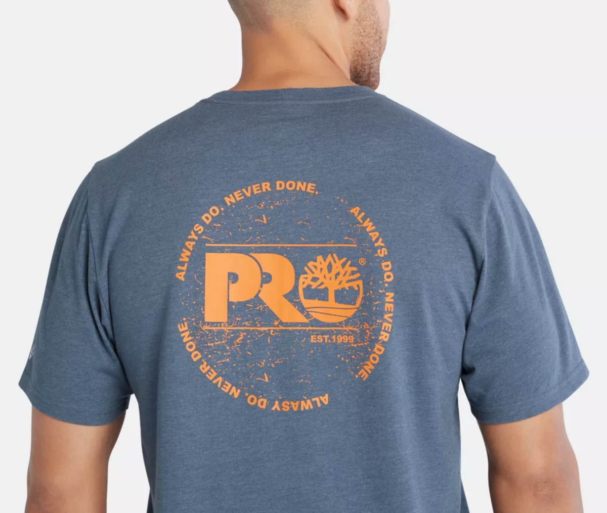 A person wearing a Timberland Pro Base Plate A.D.N.D Graphic Tee, crafted from recycled polyester, sports a blue T-shirt with an orange circular tree design and the phrases "ALWAYS DO. NEVER DONE." and "PRO EST. 1998" on the back, offering comfort with its moisture-wicking fabric.