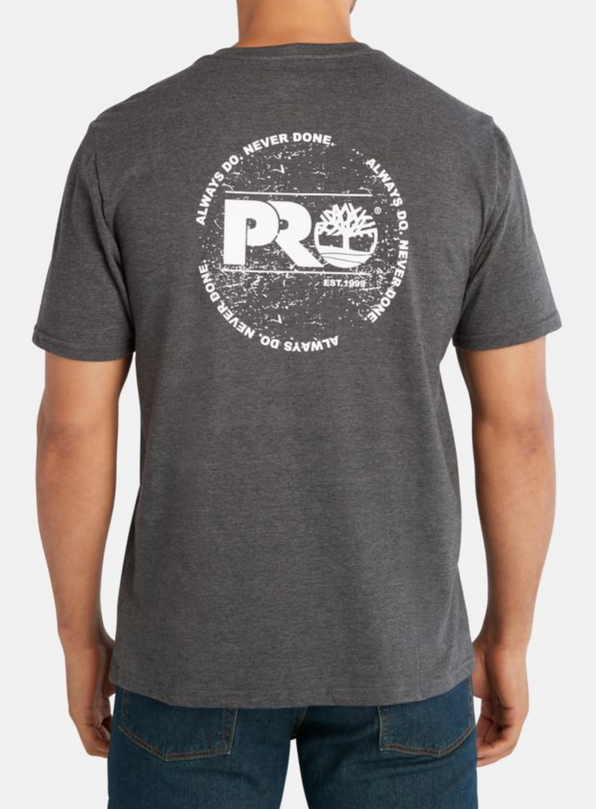 Man in a Timberland Pro Base Plate A.D.N.D Graphic Tee made from recycled polyester, showcasing a gray T-shirt with a large circular white graphic on the back, including "PRO," tree logo, "Est. 1992," and "ALWAYS DO. NEVER DONE." Paired with dark blue jeans for moisture-wicking comfort.