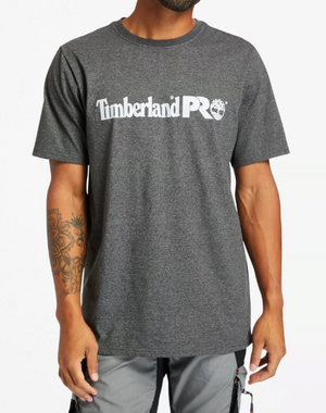 A person is wearing a gray Timberland Pro Base Plate Logo Tee with white lettering and an original fit. The wicking fabric highlights their tattooed left forearm, paired seamlessly with light gray pants against a simple white backdrop.