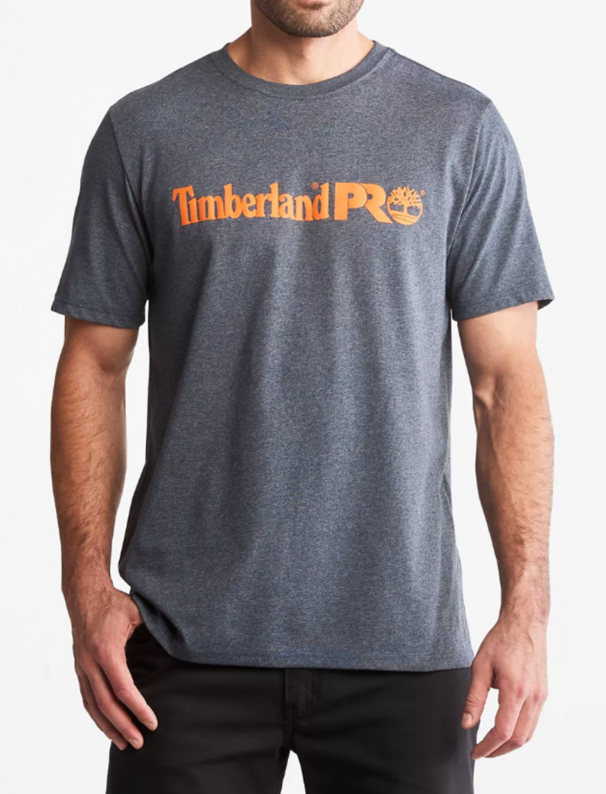 A person wears a gray Timberland Pro Base Plate Logo Tee with the signature orange "Timberland PRO" logo on the chest. Made from moisture-wicking fabric for comfort, it pairs perfectly with dark pants against a plain white background.