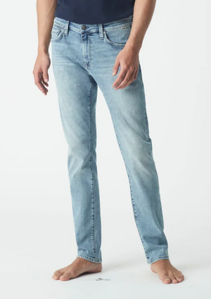 A person in Mavi Jake Slim Skinny Jeans, light blue with an ultra-stretchy, tapered fit and slightly faded five-pocket style, stands barefoot against a white background wearing a partially visible dark shirt.