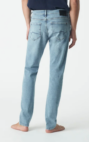 Rear view of someone in Mavi Jake Slim Skinny Jeans, light blue and ultra-stretchy with a tapered shape and classic pockets, paired with a dark shirt. They stand barefoot on a white background.