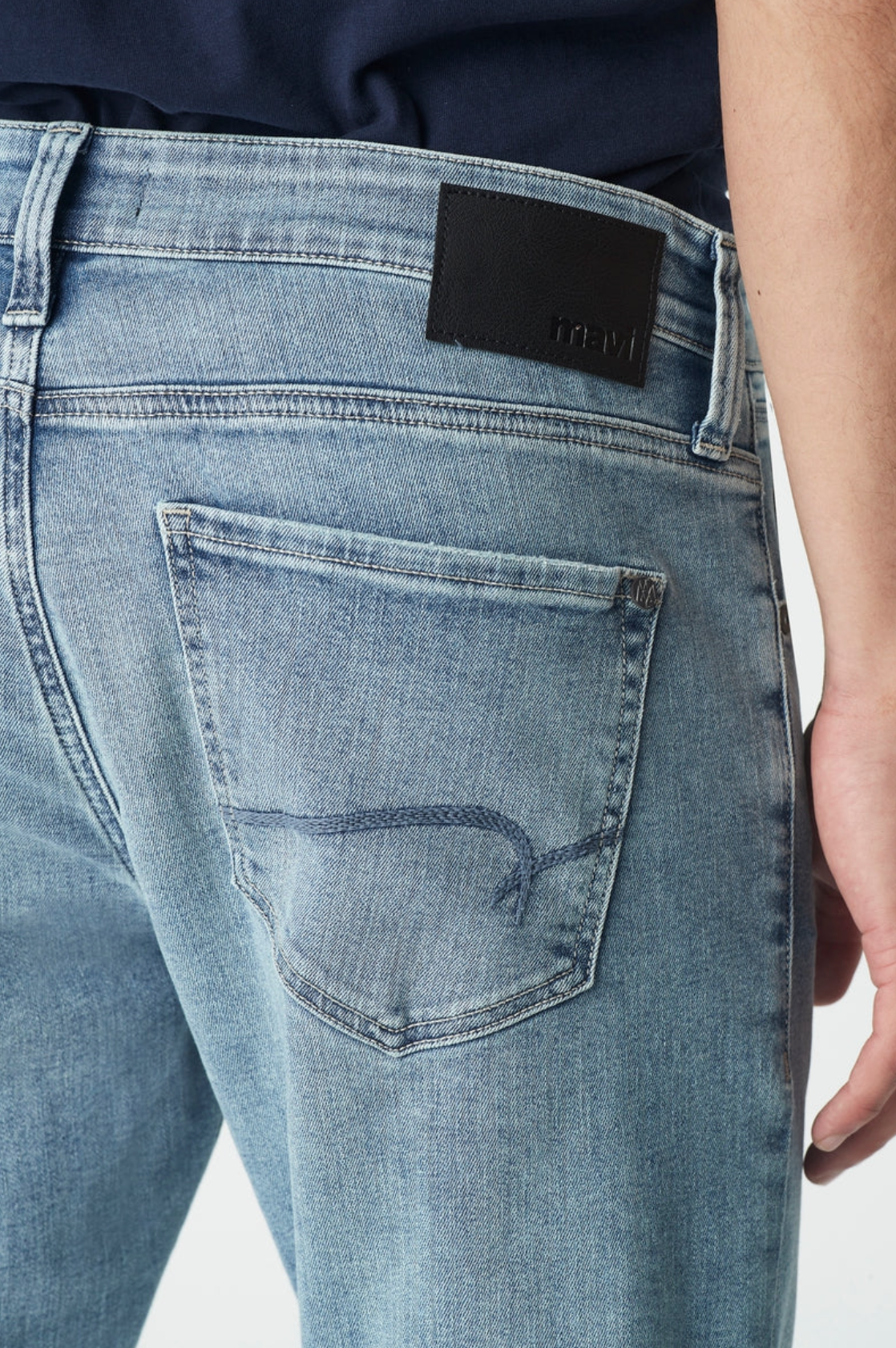 A person wearing Mavi Jake Slim Skinny Jeans, characterized by a light blue denim color and a "mavi" patch on the waistband. The ultra-stretchy fabric offers comfort, maintaining a classic five-pocket design, subtle fading, tapered shape, and is paired with a tucked-in dark shirt.