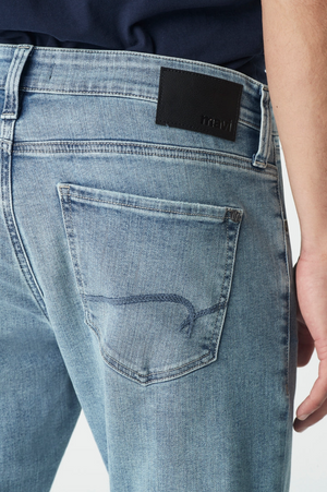 A person wearing Mavi Jake Slim Skinny Jeans, characterized by a light blue denim color and a "mavi" patch on the waistband. The ultra-stretchy fabric offers comfort, maintaining a classic five-pocket design, subtle fading, tapered shape, and is paired with a tucked-in dark shirt.