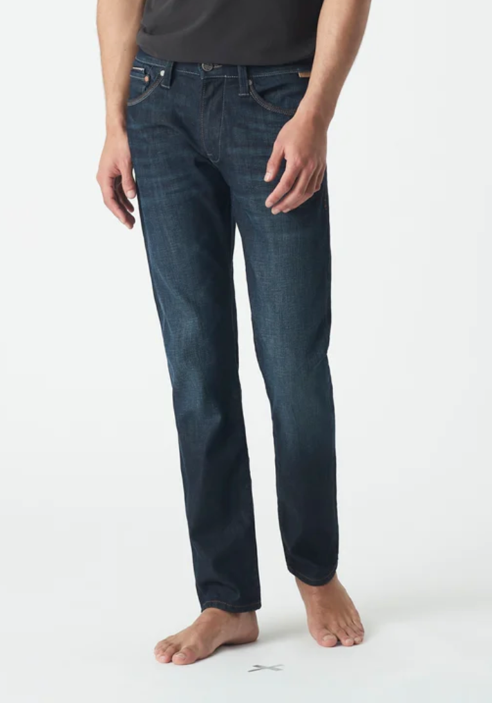 A person stands barefoot wearing Mavi Marcus Slim Straight Jeans in relaxed fit and dark blue, against a plain white background. The jeans feature visible stitching, pockets, and offer a classic slim look. The upper body is not visible.
