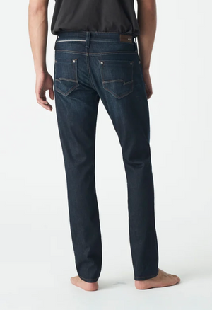 A person barefoot, facing away from the camera, wears Mavi Marcus Slim Straight Jeans in dark blue with visible stitching on the back pockets, paired with a black shirt against a plain white background.