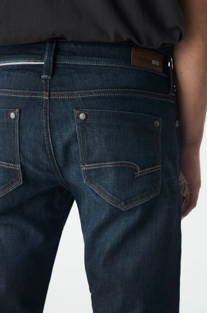 Close-up of someone in Mavi Marcus Slim Straight Jeans, featuring dark blue denim with orange stitching, metal rivets on the back pockets, and a branded leather patch on the waistband. They are facing away with part of their right arm subtly visible.