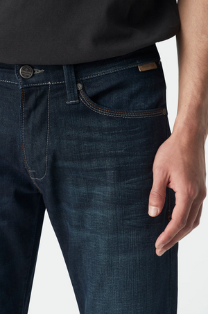 Close-up of a person in Mavi Marcus Slim Straight Jeans, dark blue and relaxed fit, with a black shirt. A hand rests on the side, highlighting the jeans' comfortable texture and precise stitching.