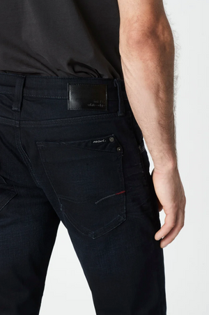 A person in dark blue Mavi Marcus Slim Straight Jeans featuring a black leather waistband patch, red stitched back pocket with a logo, wears a black shirt. Their right hand is partially visible.