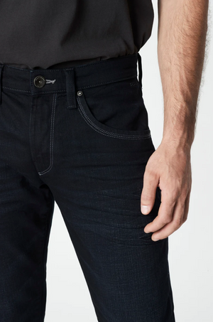 A person is pictured in the Mavi Marcus Slim Straight Jeans and a dark shirt, with their right hand near the pocket. The jeans feature a visible button and contrasting stitching, exuding a casual vibe against the plain white background.