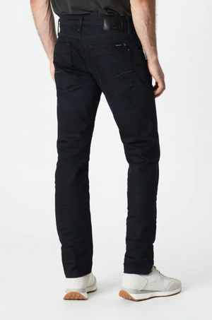A person wearing Mavi Marcus Slim Straight Jeans in black and white sneakers with tan soles stands with their back to the camera. The jeans have visible stitching and a back pocket, set against a plain white background.
