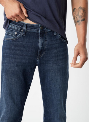 Someone is wearing a navy blue shirt and Mavi's Zach Straight Jean in dark blue. They're adjusting their shirt near the waistband with their right hand, partially revealing a tattoo of two figures on their left arm against a plain white background.