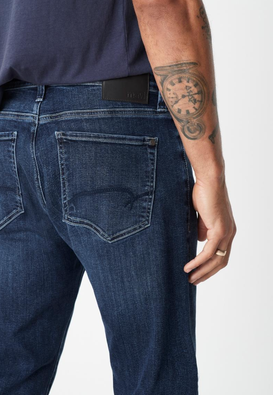 A person is seen from behind wearing Mavi Zach Straight Jeans in dark blue, crafted from organic cotton, along with a navy shirt. Their hand rests at their side, showing a tattoo on their arm; the jeans have visible back pockets and a black waistband label.