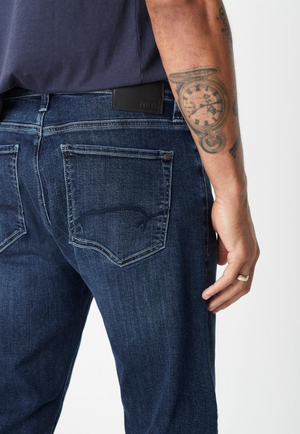 A person is seen from behind wearing Mavi Zach Straight Jeans in dark blue, crafted from organic cotton, along with a navy shirt. Their hand rests at their side, showing a tattoo on their arm; the jeans have visible back pockets and a black waistband label.