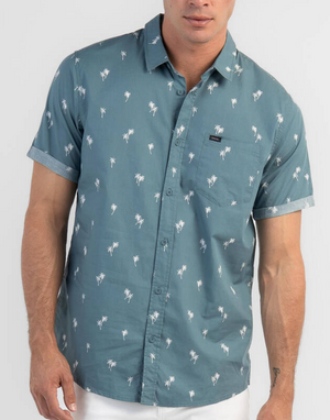 Someone is wearing a Rip Curl Paradise Palms Shirt, with short sleeves and a small palm tree pattern. This teal button-up, made from 100% cotton, has a chest pocket and is styled with white pants against a plain white backdrop.
