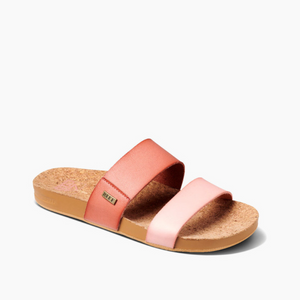 The REEF Cushion Vista Duo sandal features a cork footbed and two pink and coral straps, one with a metallic logo tag. It includes an EVA foam layer for extra comfort, set against a white background.