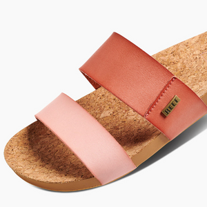 The REEF Cushion Vista Duo sandal features a stylish cork footbed, two wide straps in light pink and reddish-orange, a cushion bounce midsole for comfort, a tan textured insole, and a small metal tag with the brand name.