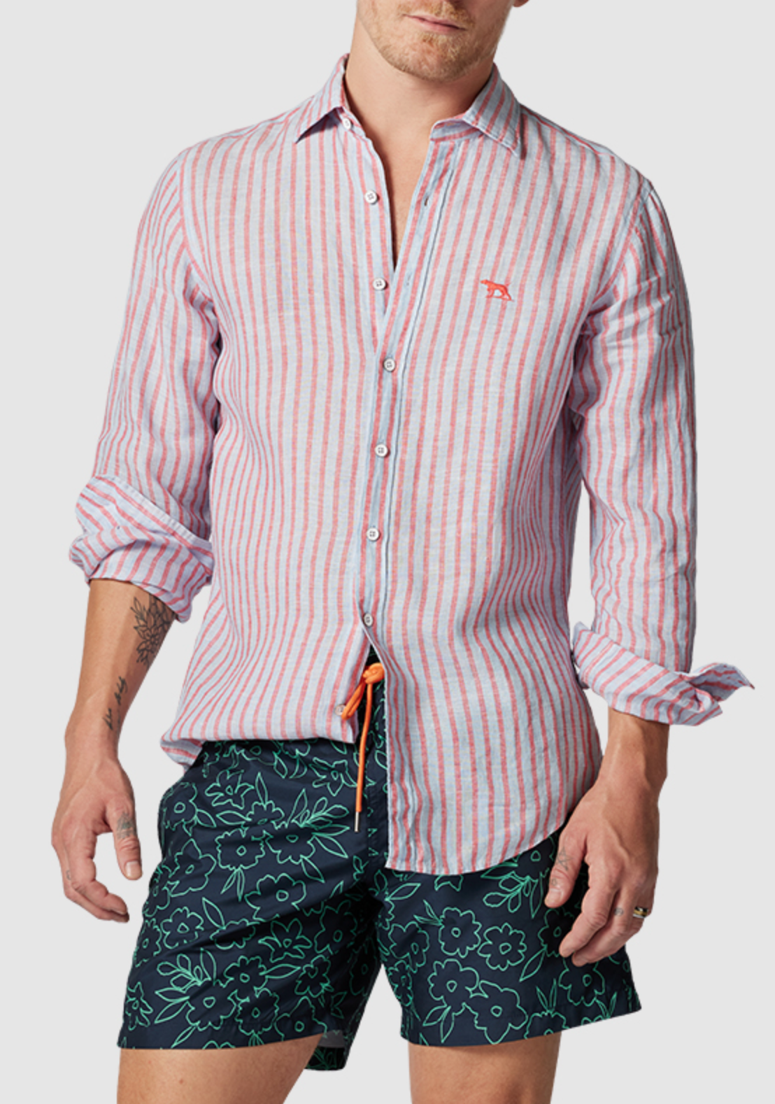 A man sports the Rodd & Gunn Mclean Park Oxford Sports Fit Shirt, a 100% linen red and white striped button-up with rolled sleeves and chest embroidery, paired with dark green floral shorts. The setting is a plain gray background.