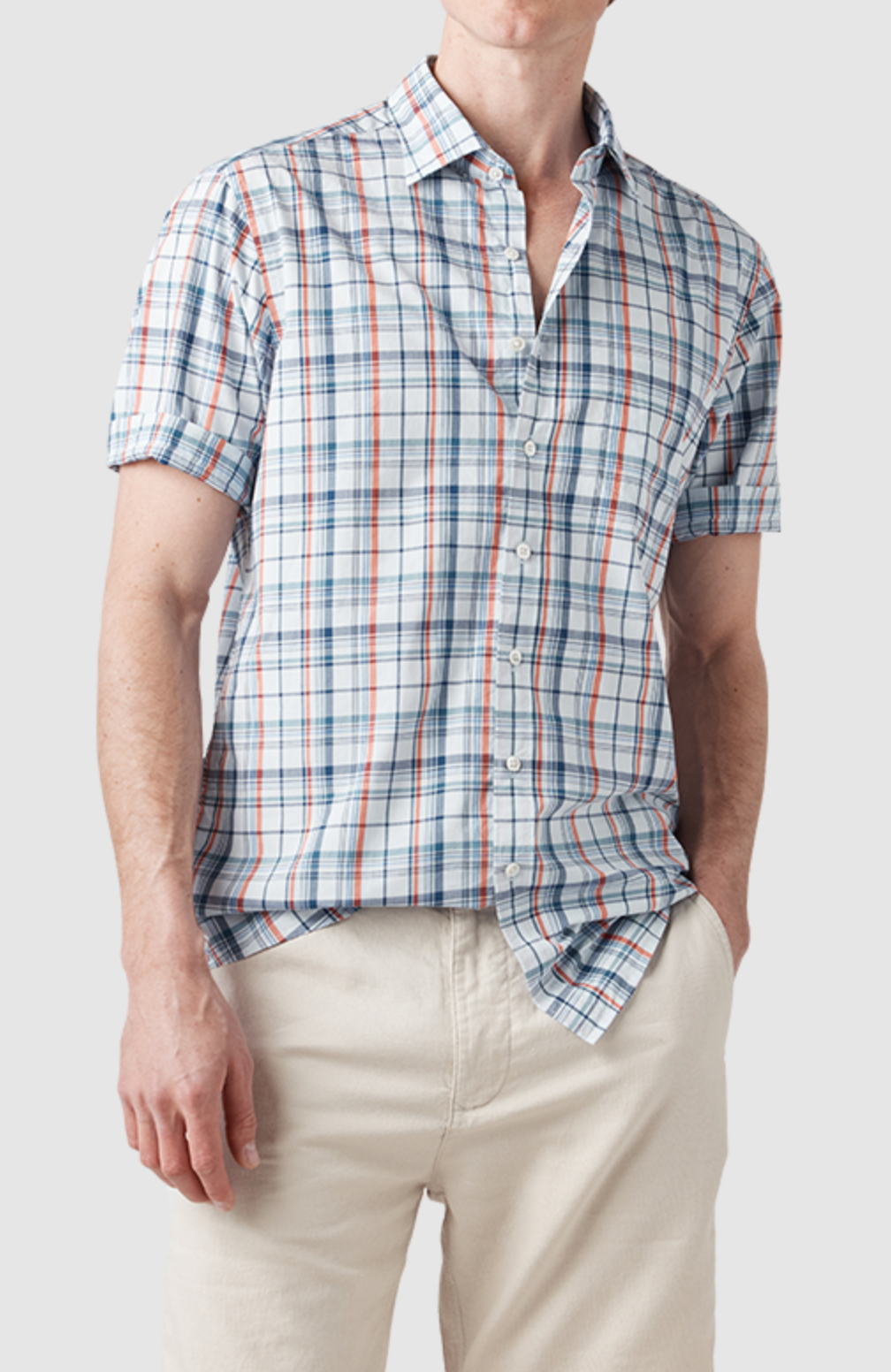 The man radiates casual style in a Rodd & Gunn Rodeo Drive Shirt, showcasing an original fit with vibrant blue and red plaid. Paired with beige pants, his left hand rests in his pocket against a plain background. Crafted from 100% cotton, this ensemble is effortlessly timeless.
