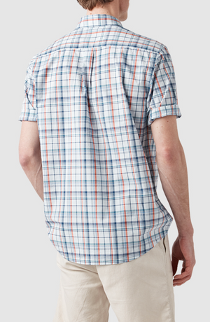 A person wearing a short-sleeved, original fit Rodd & Gunn Rodeo Drive Shirt in white and blue plaid with red accents, paired with beige pants, faces away from the camera against a gray background. The shirt is 100% cotton.