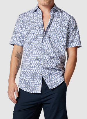 A man stands with one hand in his pocket against a gray background, wearing the Rodd & Gunn First Avenue Shirt—a stylish blue geometric-patterned, short-sleeve, 100% cotton button-up—and dark navy pants.