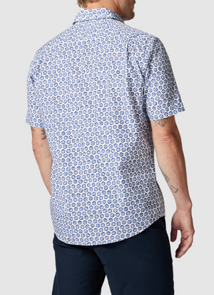 The individual sports a short-sleeved, button-up Rodd & Gunn First Avenue Shirt featuring a blue and white circular pattern and spread collar. Made of 100% cotton, it complements their dark pants. Their arm tattoos stand out against the plain gray backdrop from behind.