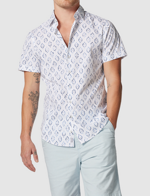 Sporting a Rodd & Gunn Penguin Place Sports Fit Shirt, someone in light blue shorts stands with one hand pocketed. The shirt's short sleeves and 100% cotton fabric showcase blue designs, enhancing the frame. A tattoo peeks out on the right arm against a plain white background.