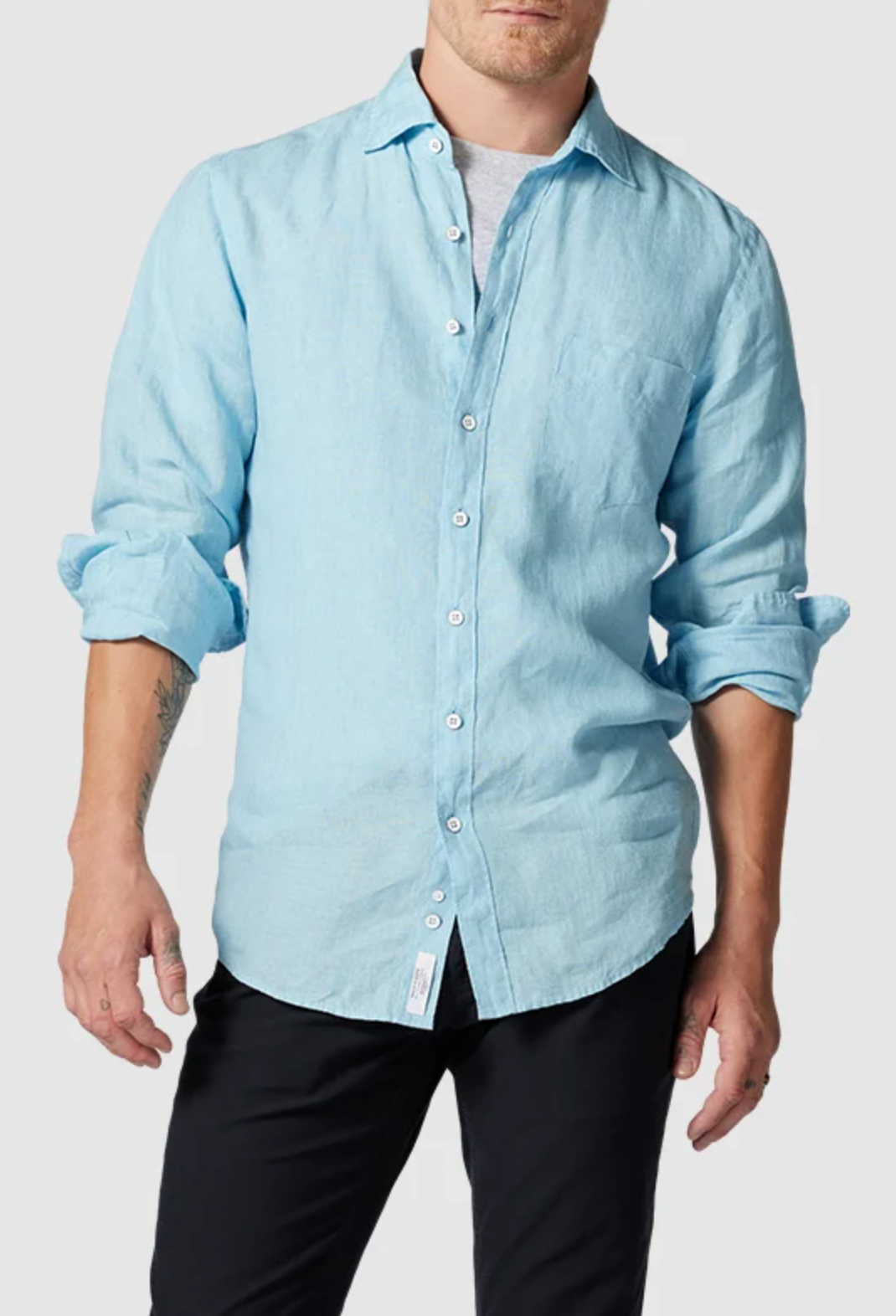 Someone wearing a light blue Rodd & Gunn Coromandel Sports Fit Shirt with sleeves rolled up and black pants. The shirt is unbuttoned at the top, showing a gray undershirt. Crafted from 100% linen, they stand against a plain light gray background.