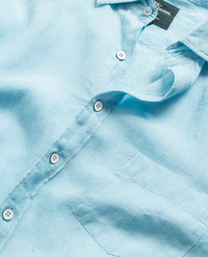 Close-up of a light blue Rodd & Gunn Coromandel Sports Fit Shirt with white buttons and a chest pocket. Laid out casually, the shirt highlights the texture of its 100% linen fabric and features a fabric label inside the collar.