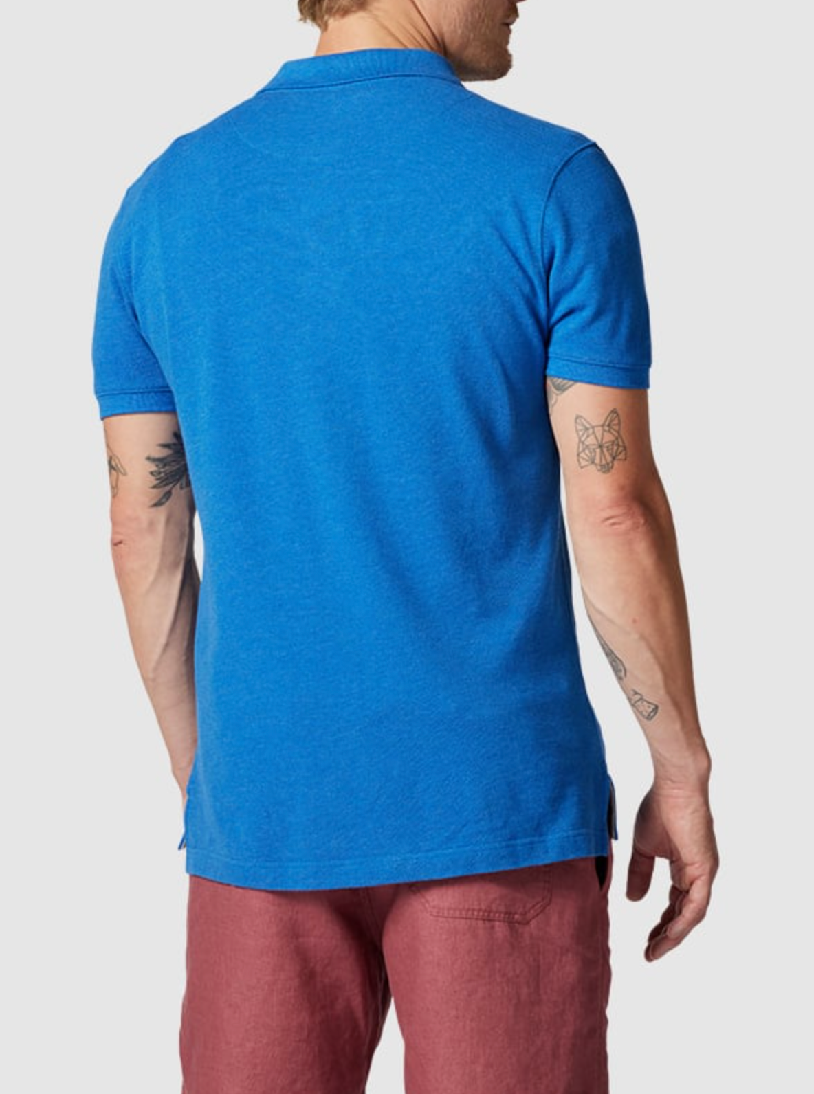 A person with short, light hair and tattooed arms, featuring a butterfly and bird design, wears the Rodd & Gunn The Gunn Polo in bright blue modern fit and pink shorts made from Australian cotton against a plain, light background.