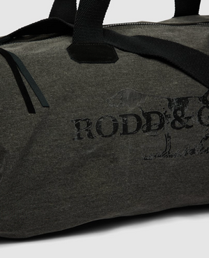 A close-up of the Rodd & Gunn Double Barrel Duffle Bag in dark grey cotton canvas with black handles, featuring a faded logo and partial dog image on its textured fabric.
