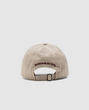 The beige Rodd & Gunn Gunn Cap, made of heavy-duty cotton twill, features the "RODD & GUNN" logo in dark red above its adjustable strap. The plain white background accentuates the cap's elegance when viewed from the back.