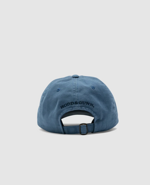The Rodd & Gunn Signature Cap, made of blue cotton twill, is shown from the back against a light gray background. The cap displays "RODD & GUNN" branding in black text and features an adjustable strap with a metal buckle.