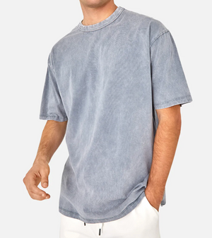 A person in a faded blue Industrie The Del Sur Acid Wash Tee, 100% cotton with a casual fit and light denim texture, stands against a plain background. One hand is in the pocket of white pants. The comfortable tee features an acid wash style, though the head isn't visible in the image.