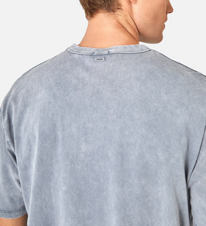 Wearing the Industrie The Del Sur Acid Wash Tee, a person is seen from behind in a blue-gray comfortable shirt, made of 100% cotton with an acid wash texture and a small neck tag. Their head is slightly turned, showing their ear.