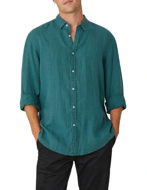 A person in a relaxed-fit, teal Industrie The Tennyson Linen Shirt with rolled sleeves and black pants stands hands-in-pockets against a plain white background.