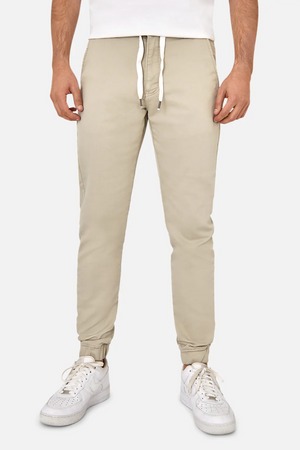 A person wearing beige Industrie The Drifter Chino Pants with an elastic waist and white sneakers stands against a plain background. The chinos have a slim fit, tapered design with elastic cuffs at the ankles.