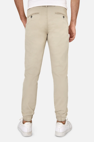 A person is shown from the back wearing Industrie The Drifter Chino Pant, slim fit in beige, and white worn-in sneakers on a plain white background. The chinos feature two buttoned back pockets, while the sneakers add a casual touch.