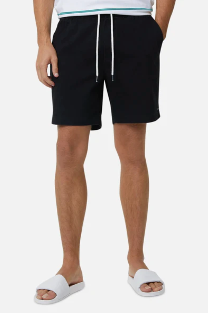 A person wearing Industrie The Belfast Short in black, featuring an elastic waist and relaxed fit, paired with a tucked-in white shirt and white open-toed slide sandals. Hands are casually in pockets against a plain white background.