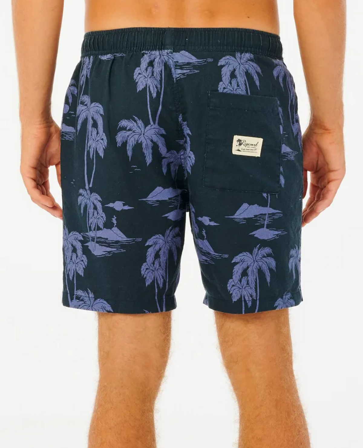 A person wears Rip Curl Mod Tropics Volley shorts in a blue tropical palm tree pattern, made from a Linen/Cotton blend. They feature an elastic waistband and a logo-patched back pocket, set against a plain white background.